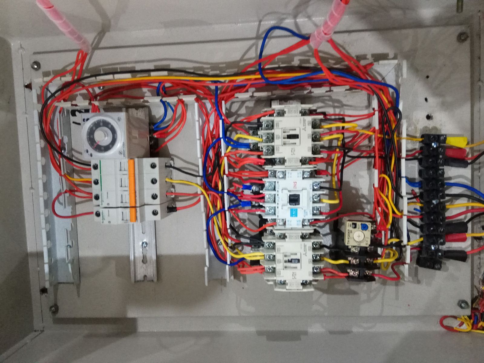 An open electrical control panel on a white wall, featuring a timer, multiple relays, and dense, colorful wiring.