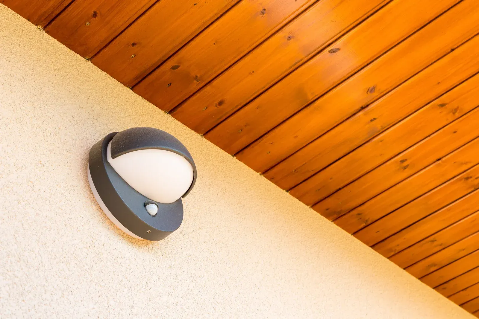 A modern round outdoor wall light with a motion sensor mounted on a beige stucco wall under a wooden ceiling.