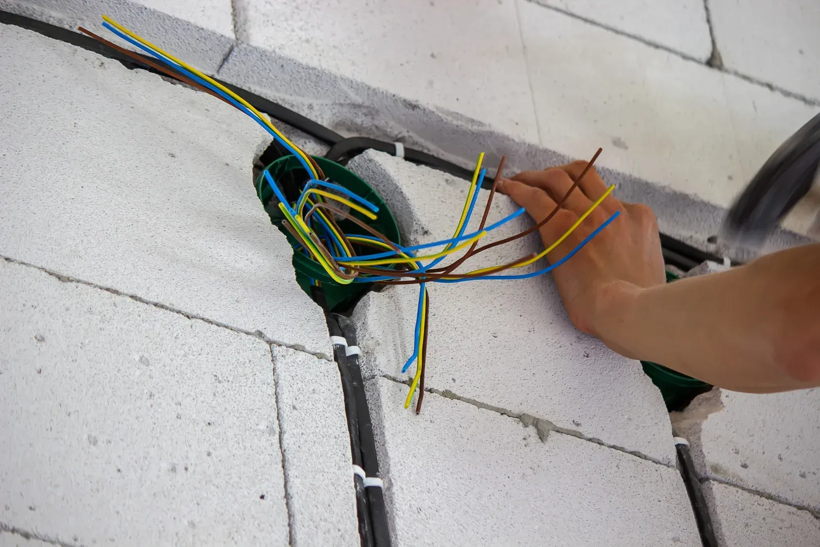 A person’s hand works on electrical wiring inside a green junction box set into a white brick wall.