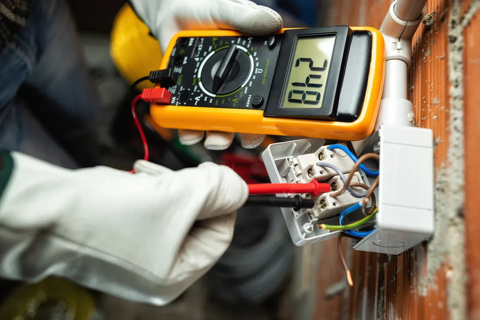 Hands wearing white work gloves use a digital multimeter to test the voltage of wires in an open electrical wall box.