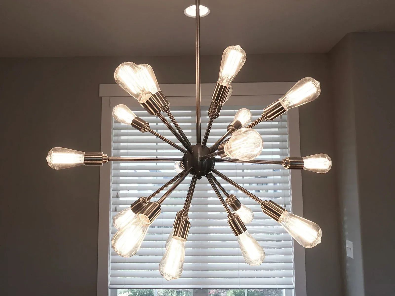 Modern brass starburst chandelier with lit bulbs hanging in front of a window with white blinds