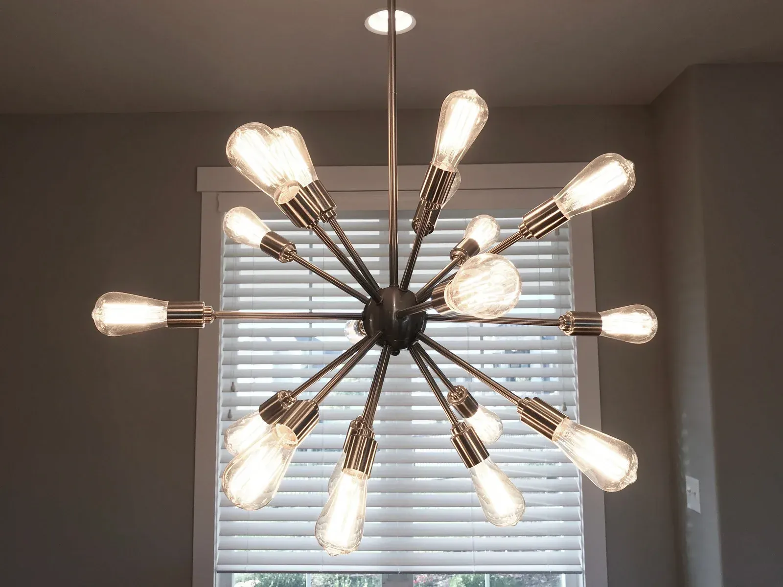 A modern, bronze-toned sputnik-style chandelier featuring multiple glowing Edison-style bulbs in front of a window.