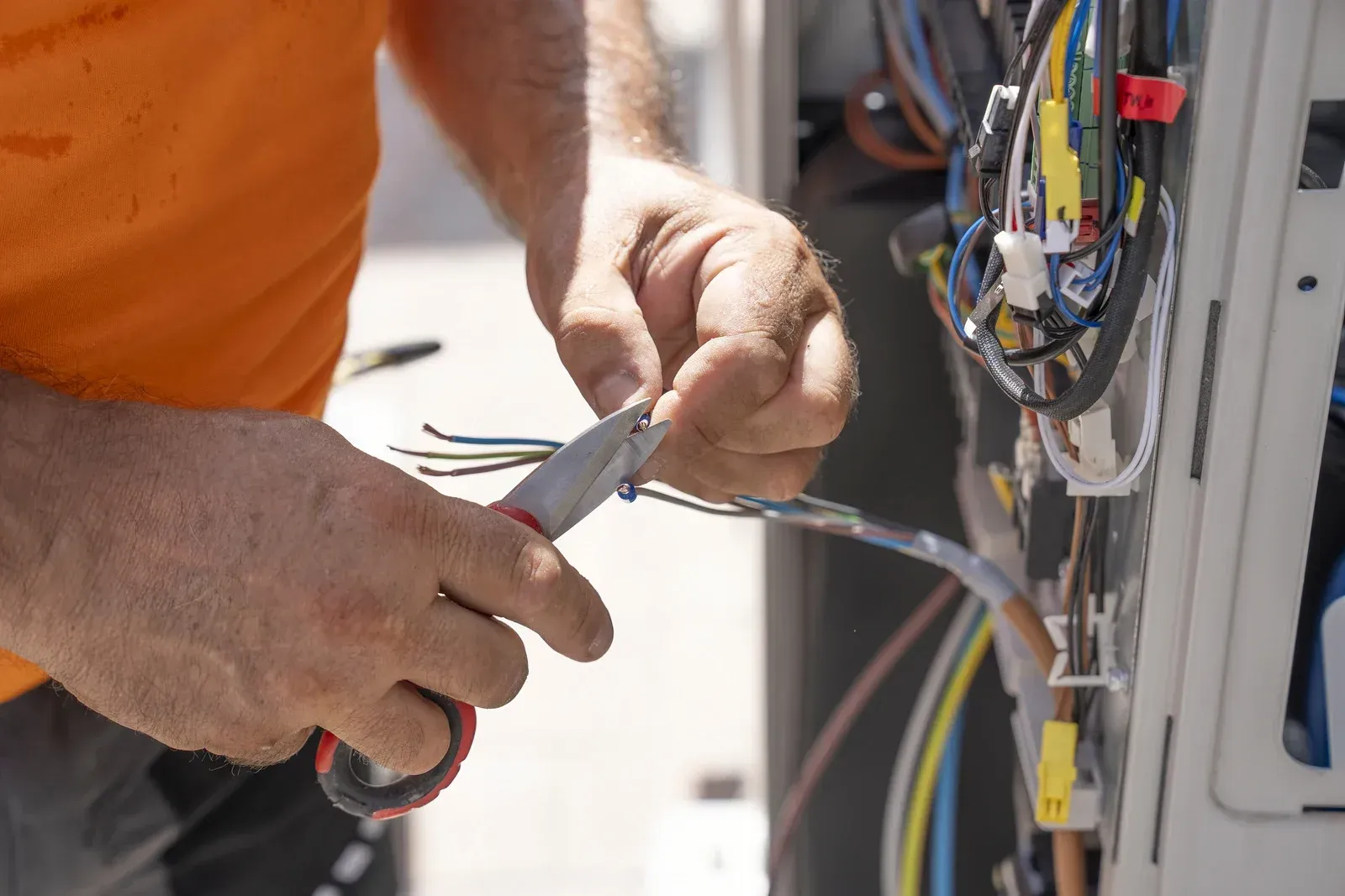 A person in an orange shirt uses wire cutters to strip the insulation off electrical wires inside an HVAC unit.