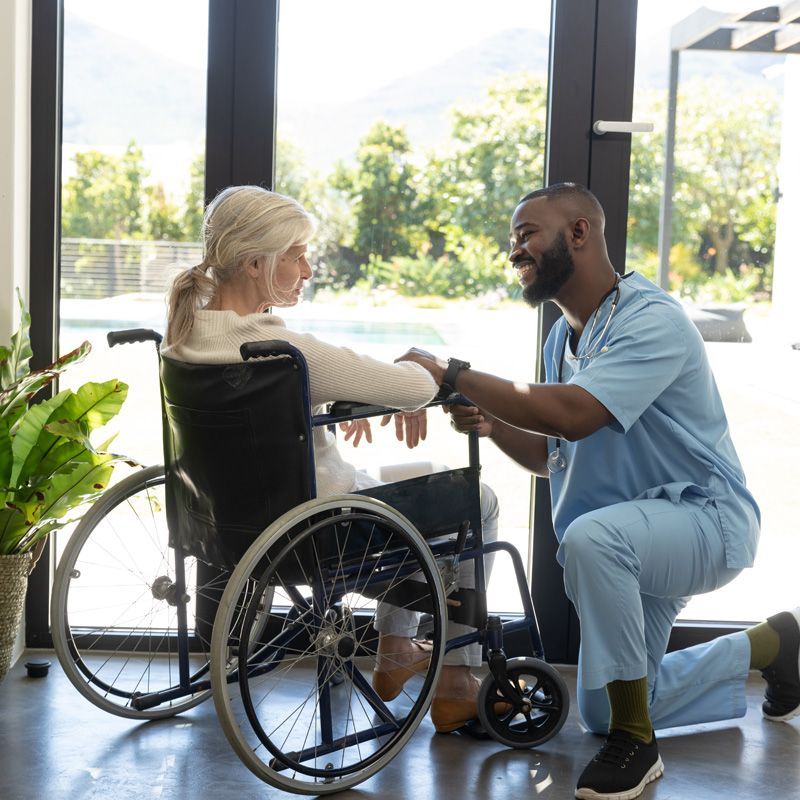 Senior woman in a wheelchair speaking with a caregiver who is checking her pulse in a bright home setting, representing compassionate senior care and mobility support.