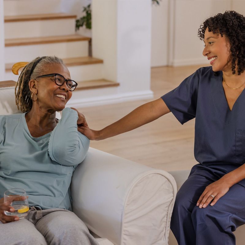 Smiling caregiver sitting with an elderly woman on a couch at home, offering companionship and support to promote senior well-being and quality of life.
