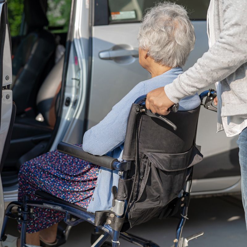 A professional caregiver assists an elderly person in a wheelchair into a medical transportation vehicle.