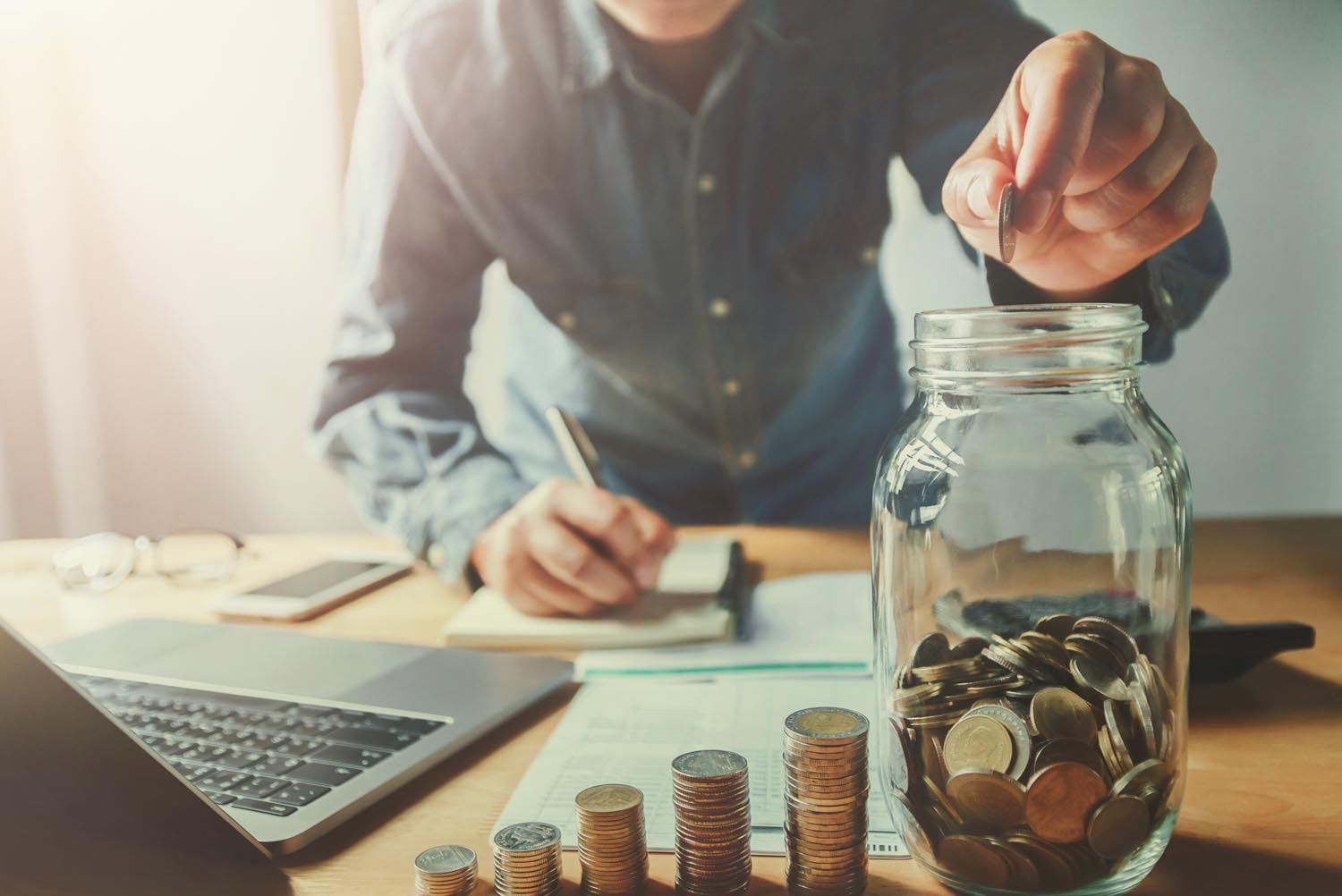 Person adding coin to jar, with stacks of coins and laptop visible, symbolizing savings.
