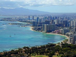 Coastal city skyline with turquoise water, beach, buildings, and mountains in the background.