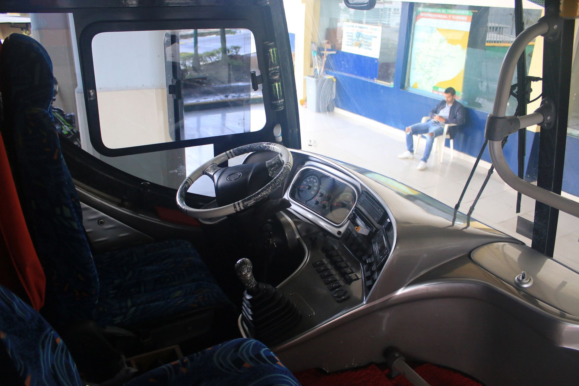Interior of a bus showing the driver's seat and a passenger waiting at the station.