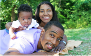 Family of three smiling outdoors: baby lying on dad's back, mom behind them.