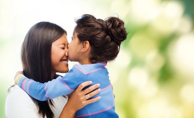 Woman smiles as a child kisses her cheek, arms wrapped around her. Green and white blurred background.