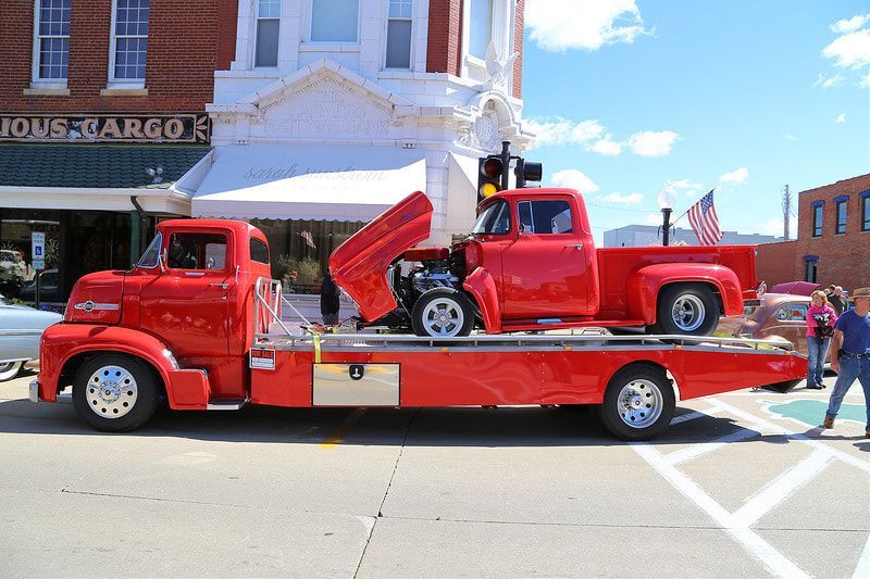 Red vintage tow truck carrying a red classic pickup truck with its hood open, street scene.