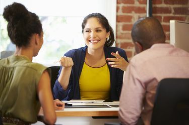 Woman in yellow top and blue blazer gestures while talking to a couple at a desk.
