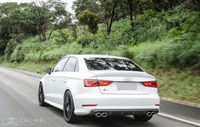 White Audi sedan driving on a road, trees in the background, exhaust pipes visible.