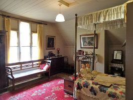 Bedroom with ornate four-poster bed, window seat, red patterned rug, and antique furniture.