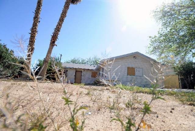 A light-colored, unfinished house under a bright sky. Foreground is dry grass and plants.