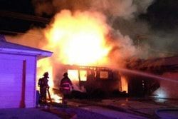 Firefighters battling a house fire at night; large flames and smoke, water being sprayed.