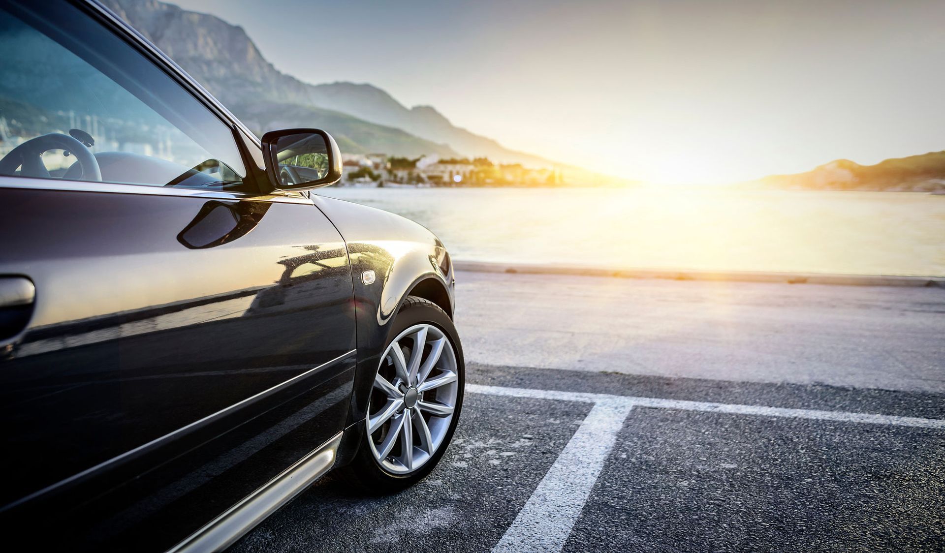 Dark car parked at a waterfront with mountains and sunlight reflecting on the water.