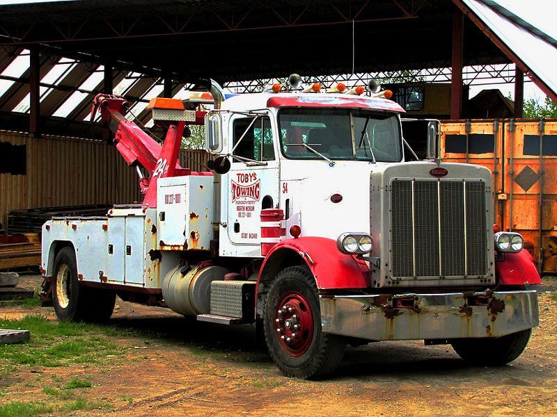 Rusty red and white tow truck with red boom, parked in a yard, under a wooden structure.