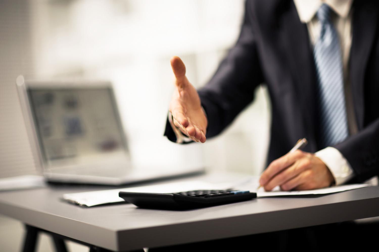 Man in suit offers handshake, standing near a desk with a laptop, calculator, and papers.