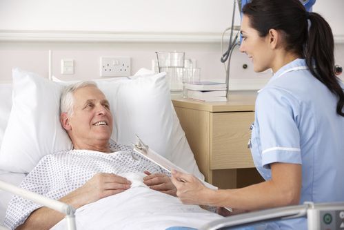 Nurse in blue scrubs talks with a patient in a hospital bed, holding a clipboard.