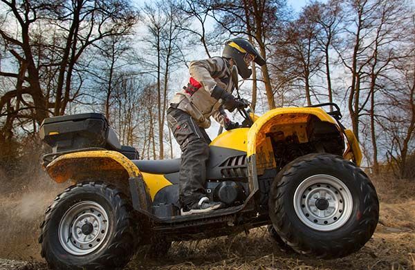 Yellow ATV driving on a dirt path in a wooded area; rider in helmet, leaning forward.