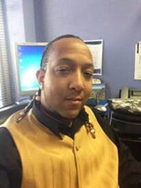 Man in a tan vest, black shirt, and earrings, sitting in front of a computer in an office.