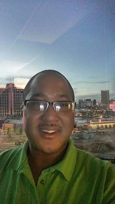 Man wearing glasses and a green shirt smiles in front of a city skyline at dusk.