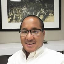 Man wearing glasses smiling, in front of a painting, wearing a white shirt.