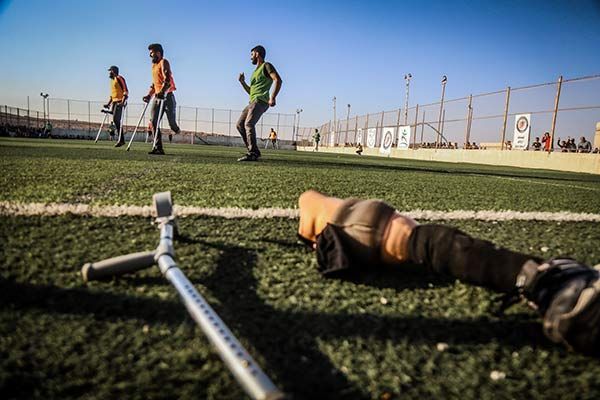 Men practice on a green soccer field using crutches and prosthetic limbs; one crutch lies in the foreground.