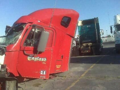 Red England semi-truck cab at a salvage yard, damaged with broken windshield, being lifted by a crane.