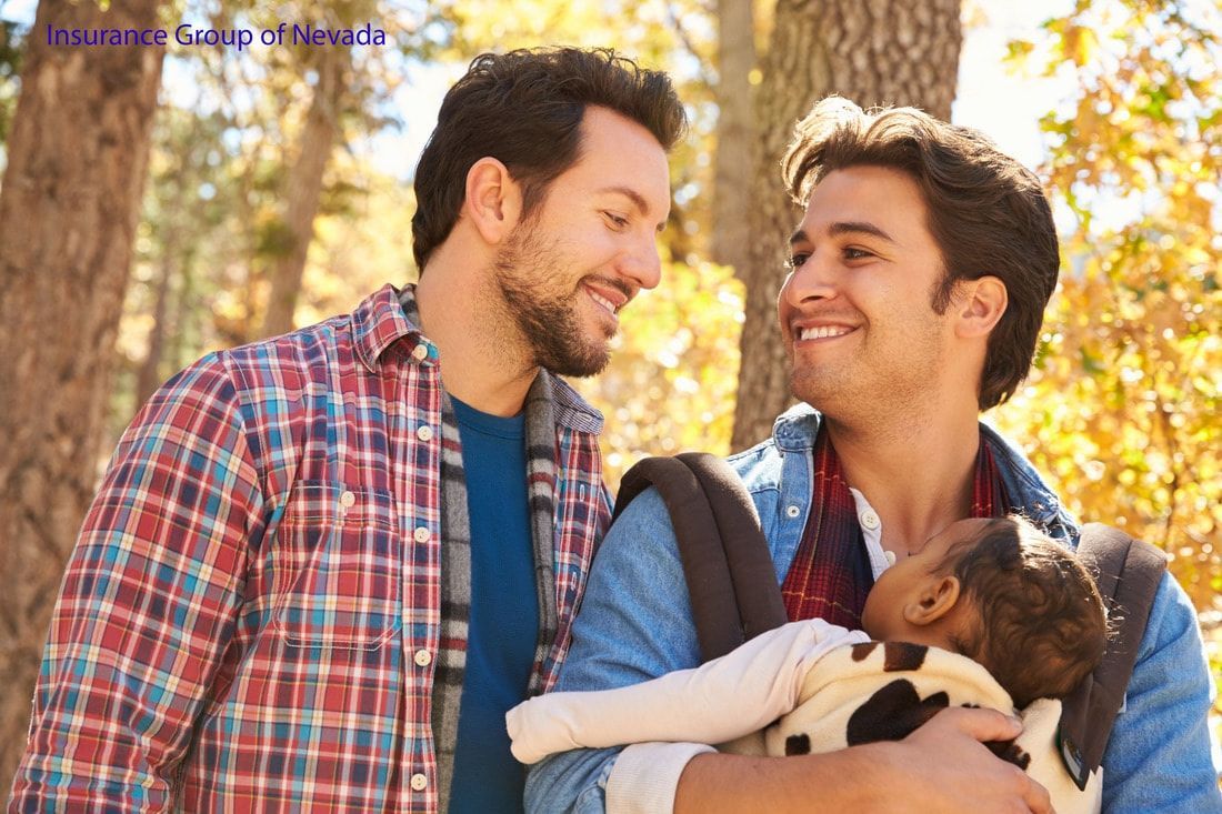 Two men smile at each other, one holding a baby in a carrier, outdoors in a park.