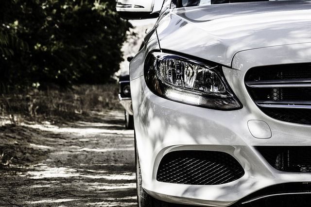 White car, close-up, parked on a dirt road. Headlight and front grill visible; another vehicle slightly behind.