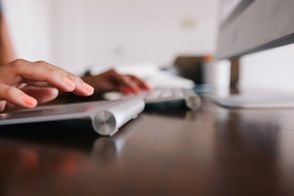Hands typing on a keyboard, with a computer monitor in the background, set on a dark wood desk.
