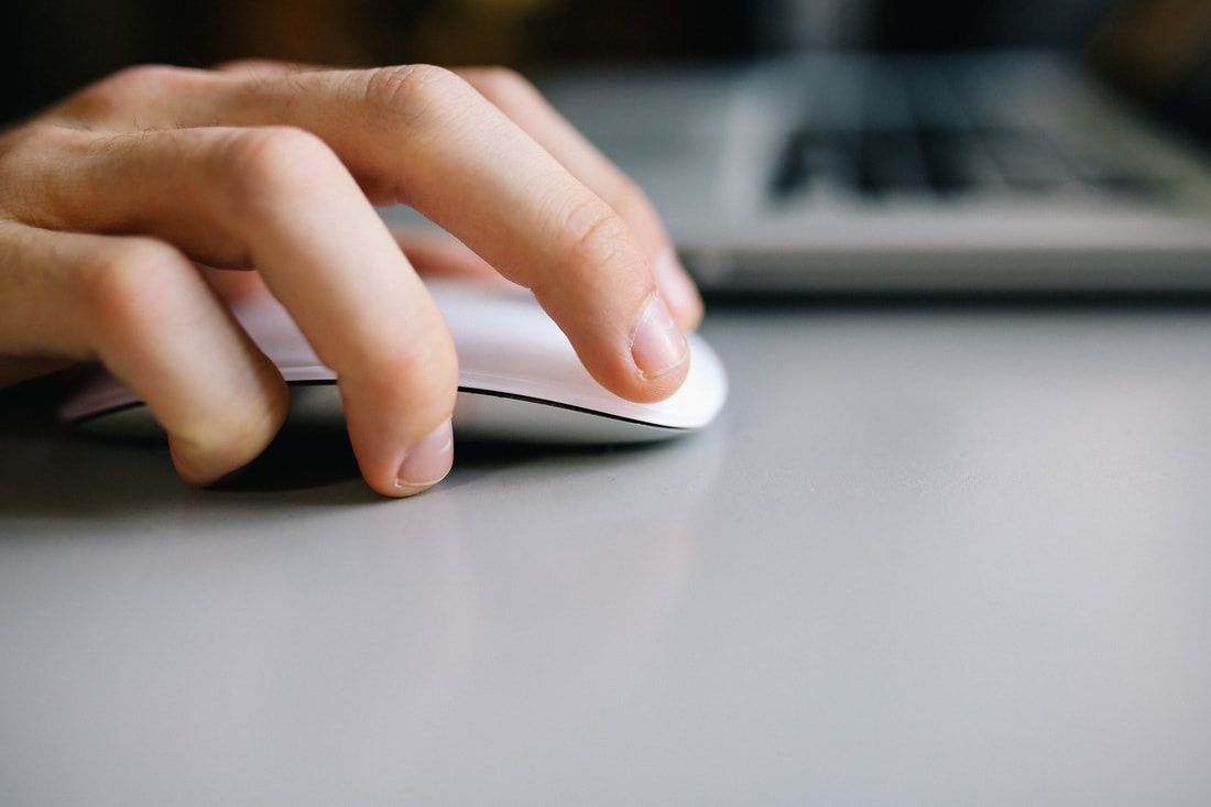 Hand operating a white computer mouse on a light gray desk, with a blurred laptop in the background.