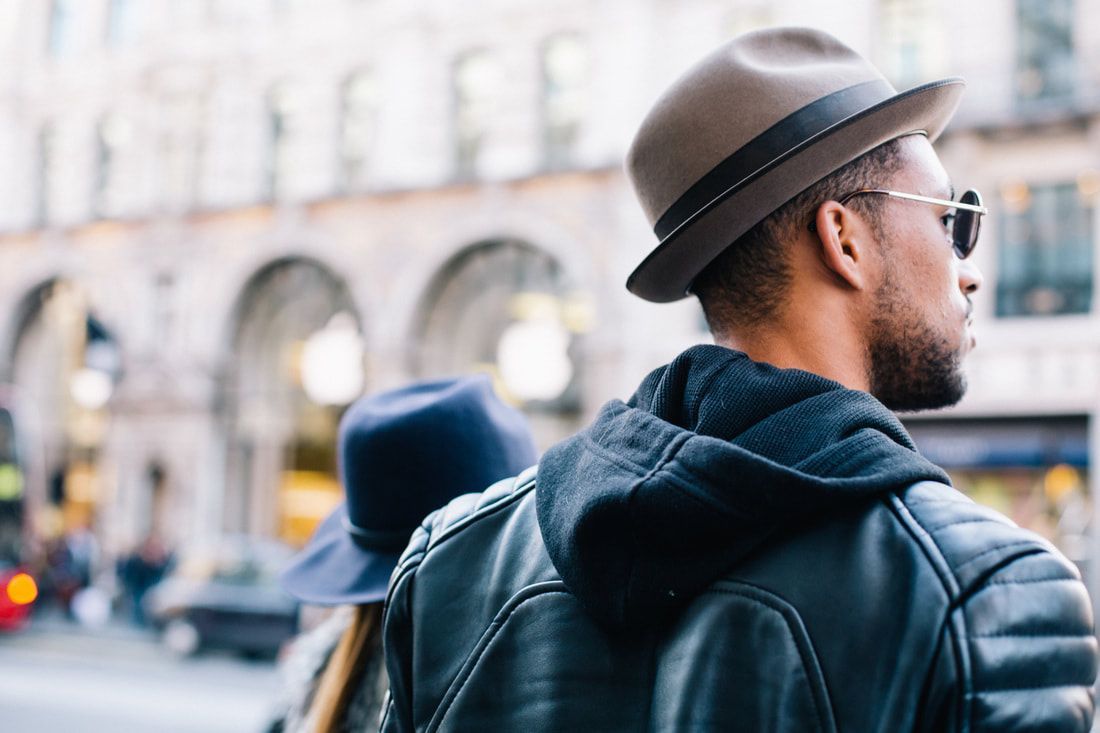Man wearing sunglasses and a fedora, leather jacket, looking off to the side, city background.