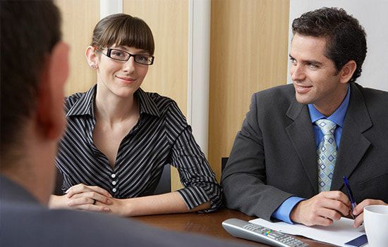 Woman and man in suits at a table, looking at someone off-camera, possibly in an interview.
