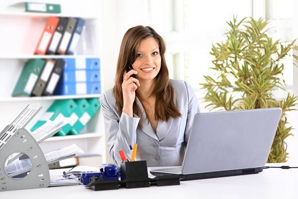 Woman in business suit smiling, on phone, at laptop in office with plant and files.