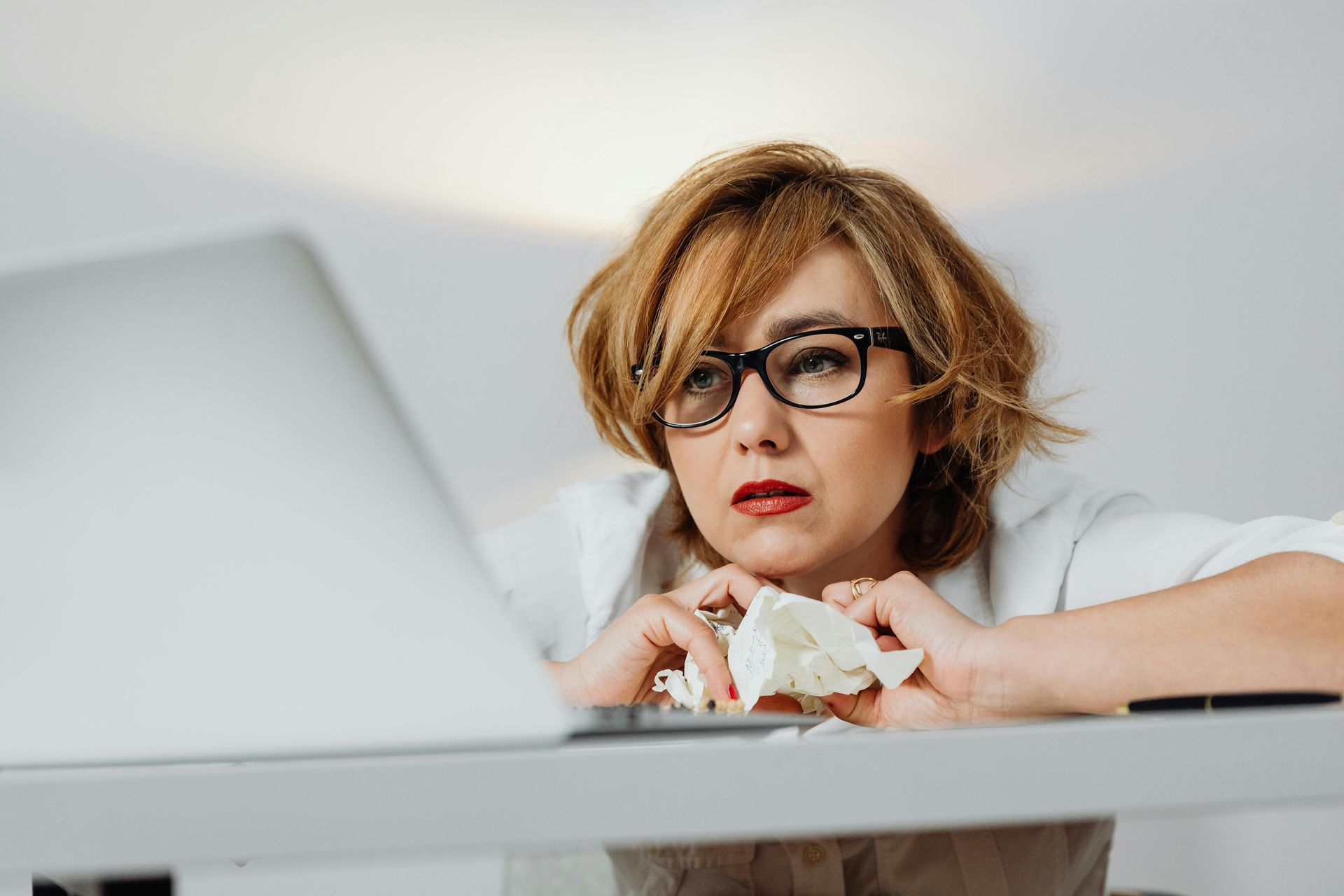 Woman with glasses looking stressed, crumpled paper in hands, at laptop on white desk.