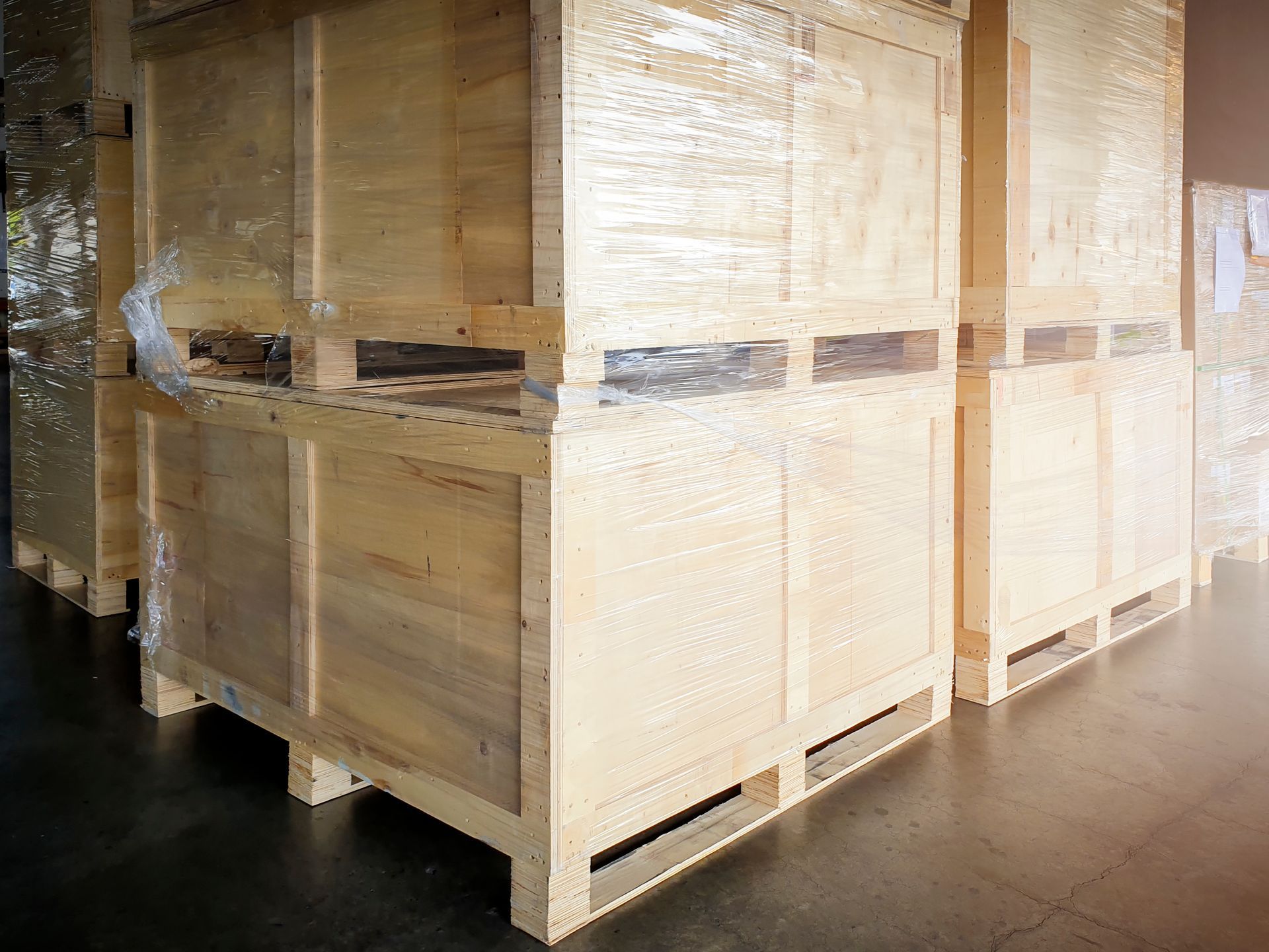 Wooden shipping crates stacked in a warehouse, wrapped in plastic and lit by warm sunlight.