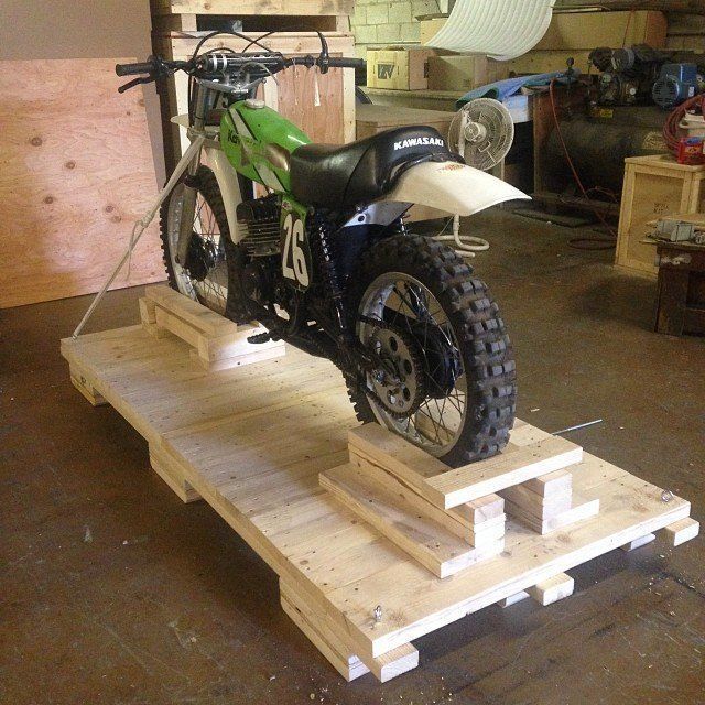 Green dirt bike on a wooden platform in a garage, secured for transport or storage.