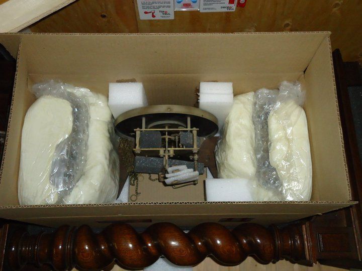 Open cardboard box with wrapped white parts and a small metal mechanism inside, resting on a wooden surface
