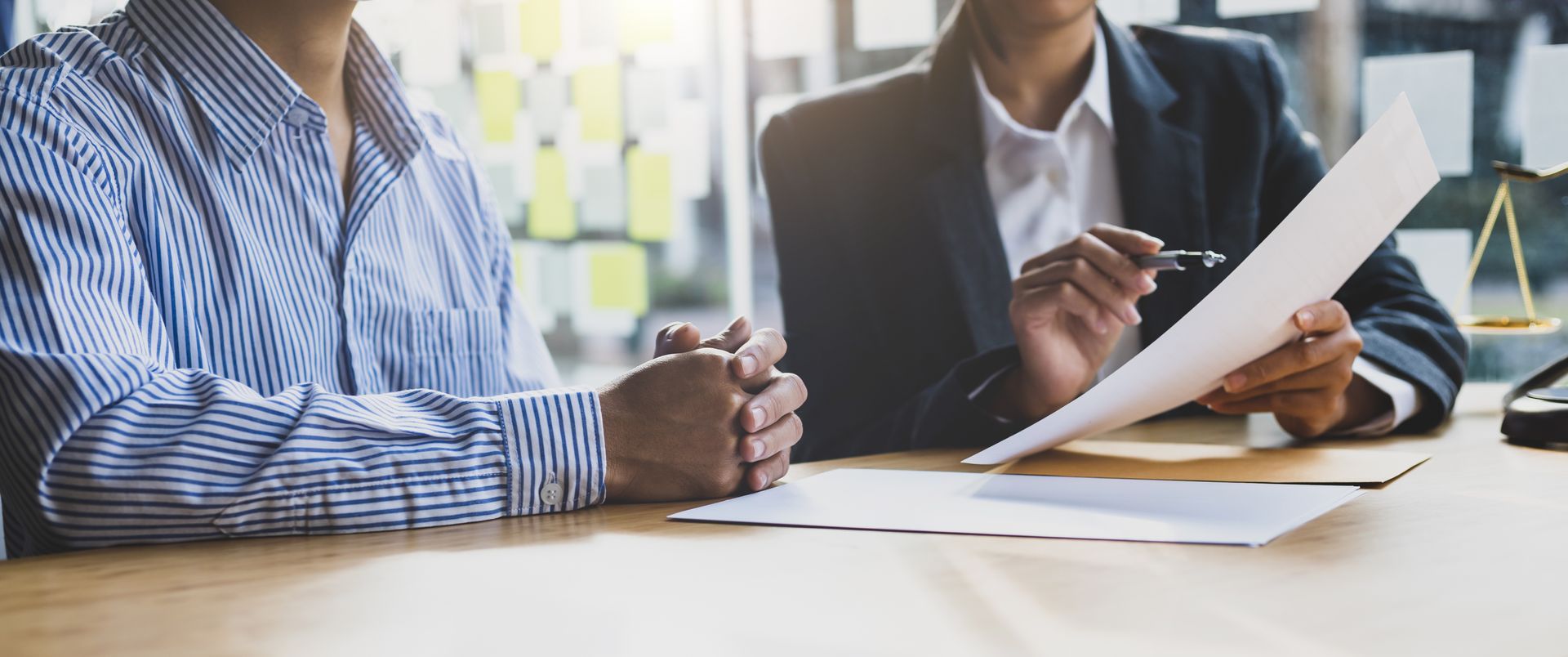 Person in striped shirt clasps hands as a suited advisor reviews paperwork by brass justice scales.