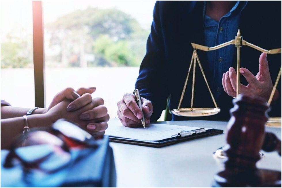 A lawyer in a suit writes on a document, with a client’s hands, a scale of justice, and a gavel visible on a desk.