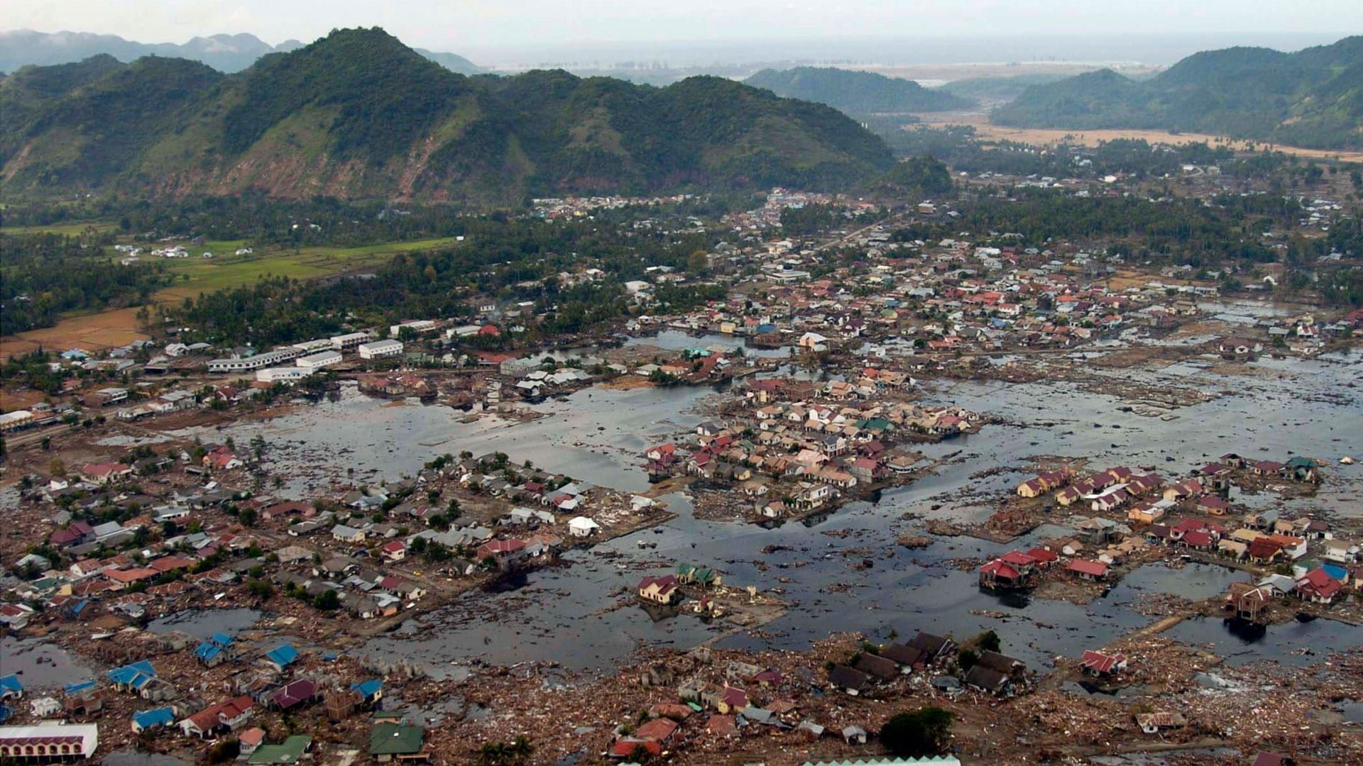 Flooded coastal town after a disaster, with damaged buildings and debris, mountains in background.