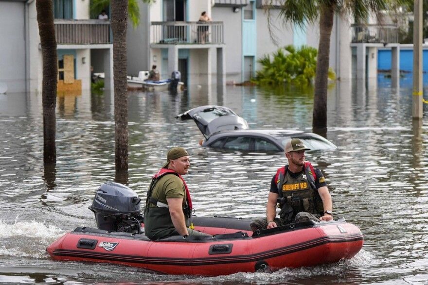 Two people in a red inflatable boat navigating floodwaters past submerged cars and buildings.