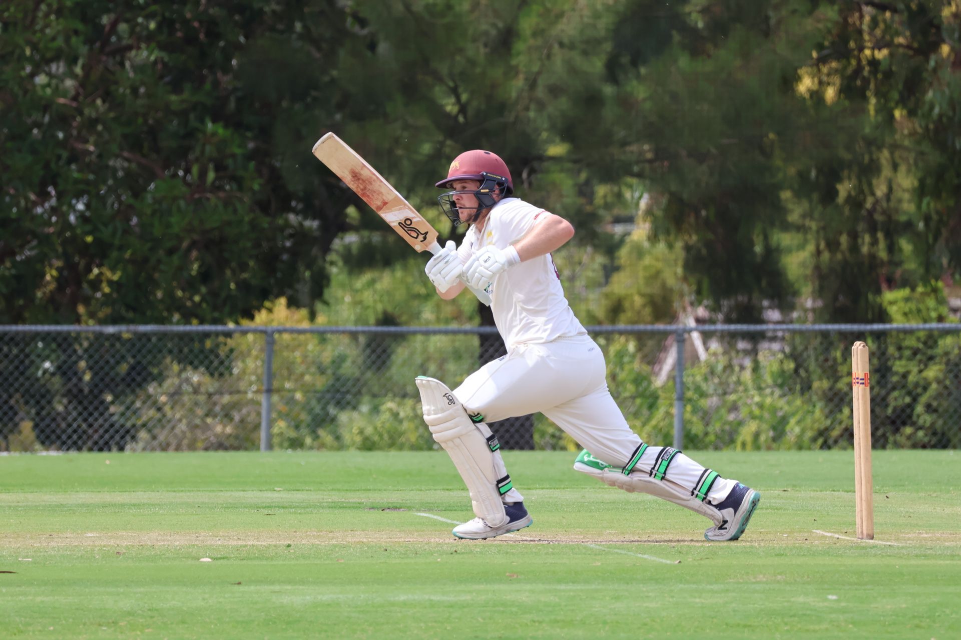 Cricketer in white uniform and maroon helmet running with bat on a grassy field, preparing to hit the ball.