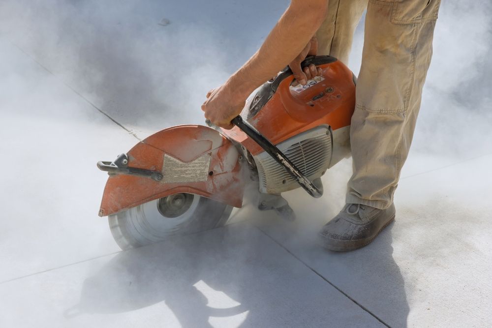 Worker using a large orange cut-off saw to cut concrete, with dust and debris around the blade.