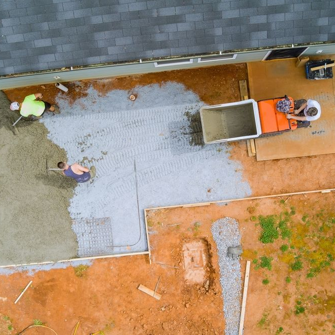 Concrete being poured for a patio by construction workers using a mixer, next to a house.