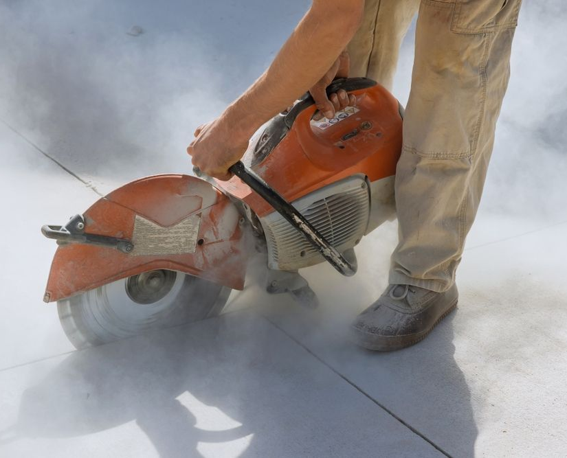 Worker using a gas-powered concrete saw cutting pavement, with dust billowing around the blade
